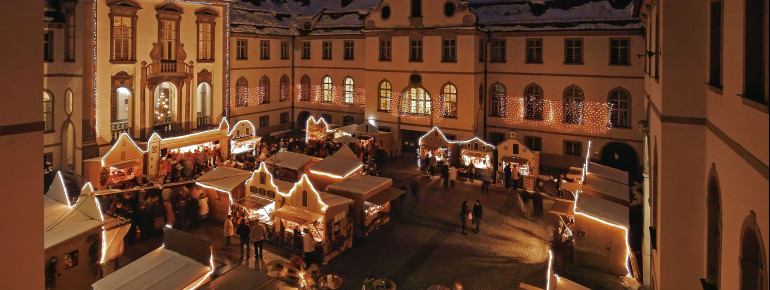 Der Adventsmarkt Füssen findet im barocken Innenhof des Klosters St. Mang statt. Im Hintergrund sieht man das beleuchtete Schloss Hohenschwangau.