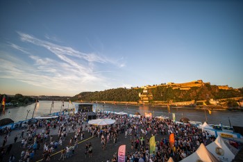 Beim Koblenzer Sommerfest feiern Besucher am Deutschen Eck, während die Festung Ehrenbreitstein im goldenen Abendlicht erstrahlt
