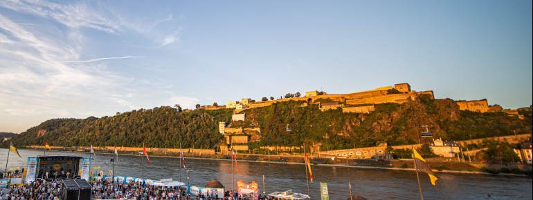 Beim Koblenzer Sommerfest feiern Besucher am Deutschen Eck, während die Festung Ehrenbreitstein im goldenen Abendlicht erstrahlt