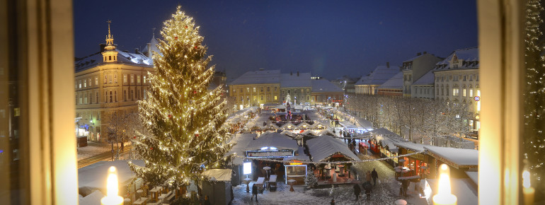 Blick vom Rathaus auf die verschneiten Buden des Klagenfurter Weihnachtsmarktes. Die große Tanne ist mit Lichterketten und Christbaumkugeln geschmückt.